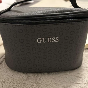 Svart necessär från Guess - Snygg och praktisk svart necessär från Guess med diskret mönster och logga i silver. Den har en dragkedja och ett bekvämt handtag för enkel transport. Insidan är rymlig och fodrad med Guess-logga.
