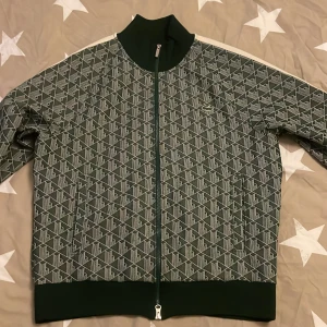  Lacoste - Säljer en snygg grön jacka från Lacoste med ett coolt geometriskt mönster. Jackan har dragkedja och är långärmad, perfekt för höst och vår. Den är i mycket bra skick och har den klassiska krokodilloggan på bröstet. Passar perfekt för en casual look!