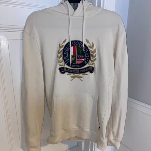 Beige hoodie med emblem - Säljer en snygg beige hoodie med ett stort emblem på bröstet. Den har en klassisk passform med långa ärmar och en praktisk magficka. Perfekt för en avslappnad stil eller för att hålla sig varm under kyliga dagar. Passar både till vardags och för lite mer avslappnade tillfällen.