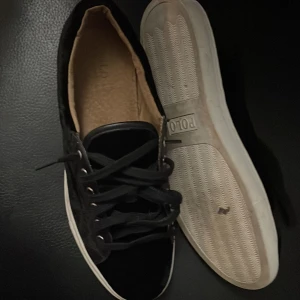 Svarta sneakers från Polo - Säljer ett par snygga svarta sneakers från Polo i bra skick. Skorna har en klassisk design med snörning och en vit sula. Perfekta för vardagsbruk och ger en stilren look. Passar till både jeans och shorts!