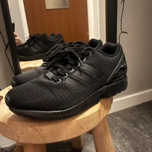Adidas ZX flux - Säljer ett par svarta Adidas sneakers i mycket bra skick. De har en stilren design med svarta ränder och snörning. Perfekta för både vardag och träning. Skorna är bekväma och har en bra passform.