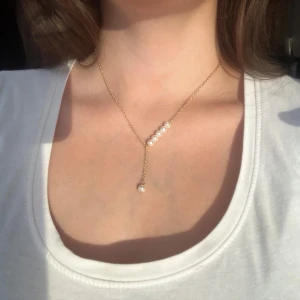 Elegant halsband med pärlor - Säljer ett stilrent och elegant halsband i guld med en rad av vita pärlor. Halsbandet har en unik design med en hängande pärla som ger en extra touch av elegans. Perfekt för både vardag och festliga tillfällen. Passar till alla outfits och ger en lyxig känsla.