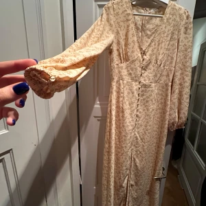 Blommig långklänning - Säljer en superfin långklänning i ljus färg med små blommor. Den har långa ärmar och en v-ringad hals. Knapparna framtill ger en söt detalj. Perfekt för vår och sommar!