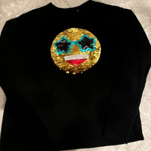 Svart tröja med paljett-emoji - Cool svart tröja med en stor emoji i paljetter på framsidan. Paljetterna kan vändas för att ändra emojins uttryck. Perfekt för att liva upp vardagslooken eller till fest! Tröjan har långa ärmar och är gjord i ett mjukt material.