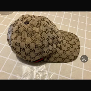 Beige keps från Gucci - Snygg beige keps från Gucci med det klassiska GG-mönstret. Kepsen har en justerbar rem baktill i brunt skinn för perfekt passform. Perfekt för att ge din outfit en lyxig touch!