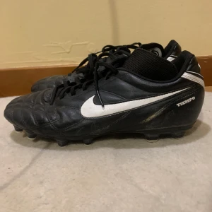 Svarta Nike Tiempo fotbollsskor - Säljer ett par svarta Nike Tiempo fotbollsskor i bra skick. Skorna har en klassisk design med vit Nike-logga och snörning. Perfekta för fotbollsträning eller match. De är bekväma och ger bra grepp på planen.