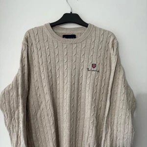 Beige stickad tröja från Bondelid - Säljer en snygg beige stickad tröja från Bondelid. Den har ett klassiskt flätmönster och ett broderat Bondelid-emblem på bröstet. Perfekt för höst och vinter, med en normal passform och långa ärmar. Supermysig och stilren!