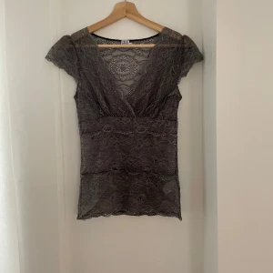 Grå spetsblus från Saint Tropez - Säljer en elegant grå spetsblus från Saint Tropez i storlek XS. Blusen har en v-ringad design och korta ärmar, perfekt för både vardag och fest. Detaljerad spets ger en romantisk touch. Passar utmärkt till både jeans och kjol för en chic look.