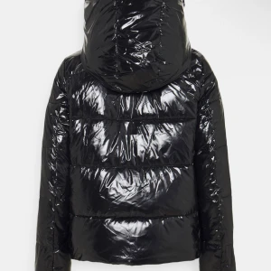 Svart glansig dunjacka från Calvin Klein - Säljer en snygg svart glansig dunjacka från Calvin Klein. Jackan har en pufferstil med en stor huva och dragkedja framtill. Perfekt för kalla vinterdagar när du vill hålla dig varm och stilren. Den är i mycket bra skick och redo för nya äventyr!