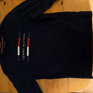 Marinblå tröja från Tommy Hilfiger - Säljer en snygg marinblå tröja från Tommy Hilfiger. Den har ett klassiskt tryck med märkesnamnet i vitt och rött på bröstet. Perfekt för en stilren look till vardags eller fest. Tröjan är i bra skick och passar perfekt till höst och vinter.
