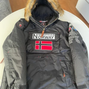 Svart vinterjacka från Geographical Norway - Säljer en svart vinterjacka från Geographical Norway med pälsfodrad huva och coola patchar. Jackan har en stor Norge-flagga på framsidan och flera detaljerade tryck på ärmarna. Perfekt för kalla vinterdagar! 🖤