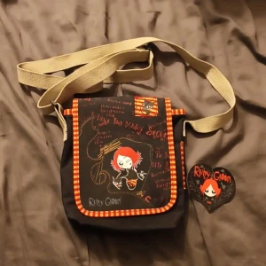 Ruby Gloom axelväska - Cool svart axelväska med motiv av Ruby Gloom. Väskan har ett justerbart beige axelband och detaljer i rött och orange randigt mönster. Perfekt för att ge en unik touch till din outfit. Den har en praktisk storlek för vardagsbruk.