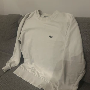 Vit sweatshirt från Lacoste - Säljer en stilren vit sweatshirt från Lacoste i mycket bra skick. Den har en klassisk design med det ikoniska krokodilmärket på bröstet. Perfekt för både vardag och avslappnade tillfällen. Storleken är inte specificerad, men den ser ut att ha en normal passform. Använd inte så mycket köpte för 1000.