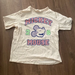 Vit/ gul  Mickey Mouse t-shirt från H&M - Säljer en vit t-shirt från H&M med ett stort tryck av Mickey Mouse i blått och rosa. T-shirten har korta ärmar och är i bomull, perfekt för en avslappnad stil. Den är i bra skick och passar både till vardags och festliga tillfällen. 🐭✨