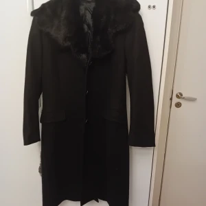 Tiger of sweden kappa - Säljer en elegant svart kappa med pälsdetaljer på kragen. Perfekt för kyliga höst- och vinterdagar. Den har en klassisk design med långa ärmar och knappar framtill. Passar både till vardags och festliga tillfällen.