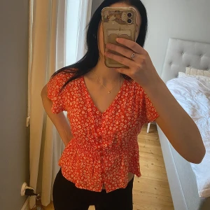 Orange blommig blus från Chiquelle - Säljer en orange blommig blus från Chiquelle. Den har korta ärmar och en söt volangdetalj i midjan. Den är stretchig i midjan och har knappar som går att öppna. Är i strl L men sitter som en S/M. 💗