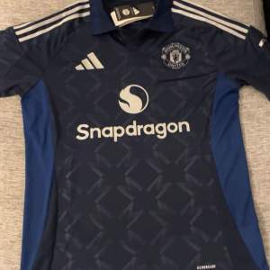 Säljer en snygg blå Manchester United fotbollströja från Adidas. Tröjan har korta ärmar och ett coolt mönster över hela framsidan. Den har vita detaljer och loggan för Snapdragon på bröstet. Perfekt för alla United-fans! 💙tröjan är Äkat och helt ny oanvänd.