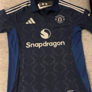 Blå Manchester United fotbollströja - Säljer en snygg blå Manchester United fotbollströja från Adidas. Tröjan har korta ärmar och ett coolt mönster över hela framsidan. Den har vita detaljer och loggan för Snapdragon på bröstet. Perfekt för alla United-fans! 💙tröjan är Äkat och helt ny oanvänd.
