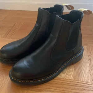 Svarta Dr. Martens boots, Chelsea - Säljer ett par klassiska svarta Dr. Martens boots i skinn. De har den ikoniska gula sömmen och är i mycket bra skick. Perfekta för både höst och vinter med en robust sula som ger bra grepp. Passar till både vardag och fest! Modell 2976 Black Ambassador. Köpta I London på Dr Martens butik. 