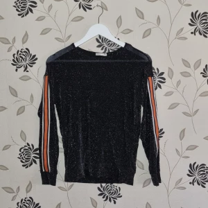 Svart glittrig tröja med ränder på ärmarna, Nly Trend, Strl: S/36 - Säljer en svart glittrig tröja i storlek S. Den har långa ärmar med coola ränder i orange och vitt längs sidorna. Perfekt för en festlig kväll eller när du vill sticka ut lite extra. Tröjan är i bra skick och redo för nya äventyr! ✨