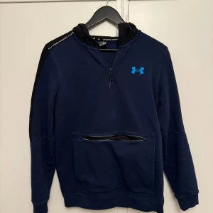 Mörkblå hoodie från Under Armour - Säljer en snygg mörkblå hoodie från Under Armour i storlek S. Den har en halv dragkedja och en praktisk ficka framtill. Perfekt för träning eller vardagsbruk. Huvan har en stilren svart insida och loggan syns tydligt på bröstet. Passar perfekt för höst och vår!