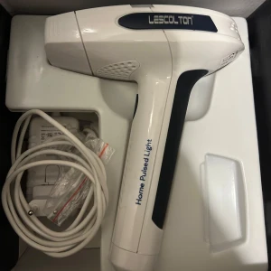 Hårborttagning maskin - Lescolton Home Pulsed Light-enhet för hårborttagning. Den är vit med svarta detaljer och kommer med en strömadapter och utbytbara lampor. Perfekt för hemmabruk för att enkelt ta bort oönskat hår.