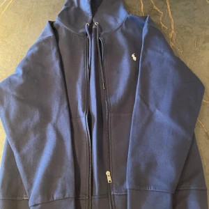 Mörkblå hoodie från Ralph Lauren - Säljer en snygg mörkblå hoodie från Ralph Lauren. Den har en dragkedja framtill och en liten logga på bröstet. Perfekt för höst och vår, och den är i mycket bra skick. Passar både till vardags och lite mer avslappnade tillfällen. Skön och stilren design!