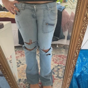 Ljusa jeans med slitningar - Säljer ett par ljusa jeans med coola slitningar på knäna och en unik ficka med dragkedja. De har en bootcut-stil och är lågmidjade!
