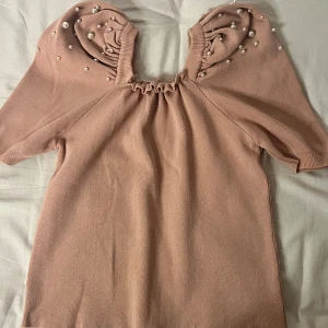 Rosa topp med pärlor - Superfin rosa stickad topp med korta ärmar och söta pärldetaljer på axlarna. Perfekt för både vardag och fest! Den har en rynkad halslinje som ger en unik touch. Passar perfekt till jeans eller kjol för en chic look.
