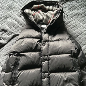 Svart pufferjacka från Burberry - Säljer en snygg och varm svart pufferjacka från Burberry. Jackan har en quiltad design med dragkedja och en mysig fodrad insida med ett klassiskt rutigt mönster. Perfekt för kalla vinterdagar! Går även att ta bort ärmarna så den blir en väst. BARA SERIÖSA❤️