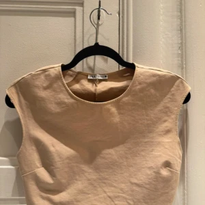 Beige ärmlös topp - Säljer en stilren beige ärmlös topp. Modellen är kort. Perfekt för sommardagar eller som ett basplagg i garderoben. Toppen har en enkel och elegant design med rund halsringning.
