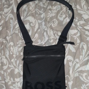 Svart axelväska från Boss säljer den för den kommer inte till användning  - Säljer en stilren svart axelväska från Boss. Väskan har en justerbar rem och en praktisk dragkedja framtill. Perfekt för att bära med sig det nödvändigaste på ett snyggt sätt.