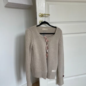 Beige stickad kofta från Odd Molly - Säljer en superfin beige stickad kofta från Odd Molly. Den har långa ärmar och en söt knytning framtill. Det finns en broderad kompassdetalj med texten 'LOVE' på ryggen, vilket ger den en unik touch. Perfekt för höst och vinter när du vill vara både varm och stilren. 🧥✨