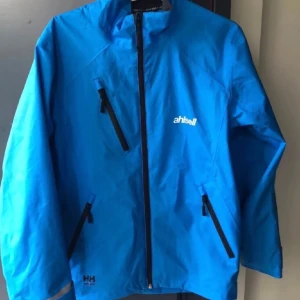 Blå vindjacka från Helly Hansen - Säljer en snygg blå vindjacka från Helly Hansen. Jackan har en dragkedja framtill och flera praktiska fickor med dragkedjor. Perfekt för blåsiga dagar och outdoor-äventyr. Den är i storlek XS och passar bra för dig som gillar en tight passform.