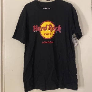 Svart Hard Rock Cafe T-shirt från London - Säljer en klassisk svart T-shirt från Hard Rock Cafe i London. Den har det ikoniska Hard Rock Cafe-loggan i rött och gult på framsidan. T-shirten är i nyskick och har kvar prislapp. kvitto finns på bild 4 & priset i svenska kronor på Bild 5.