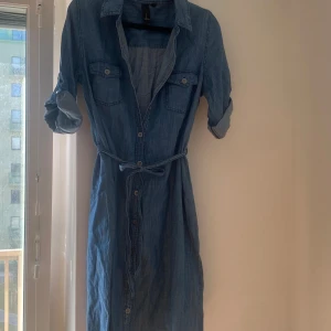 Jeansklänning med knytband - Säljer en snygg jeansklänning i blått med knytband i midjan. Klänningen har långa ärmar som kan rullas upp och knäppas med knappar framtill. Perfekt för en avslappnad dag eller en sommarfest!