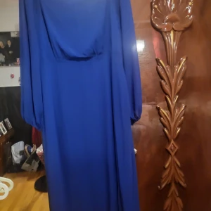 Blå långklänning med långa ärmar - Säljer en elegant blå långklänning med långa ärmar. Klänningen har en draperad design och är perfekt för formella tillfällen. Materialet ser ut att vara lätt och flödande, vilket ger en vacker siluett.