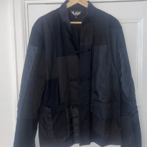 Svart jacka från Comme des Garçons - Säljer en svart jacka från Comme des Garçons, tillverkad i Japan. Jackan har en unik design med olika material som bomull, ull, nylon och polyester. Den har en stilren look med knäppning framtill och är perfekt för både höst och vår.