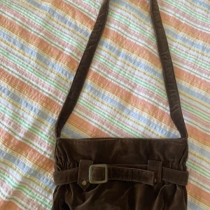 Brun axelväska från Bruuns Bazaar - Säljer en snygg brun axelväska från Bruuns Bazaar. Väskan är i mocka och har en cool bältesdetalj på framsidan. Insidan är fodrad med ett blommigt mönster i rosa och orange, och det finns en praktisk dragkedja för extra säkerhet. Perfekt för höst och vår!