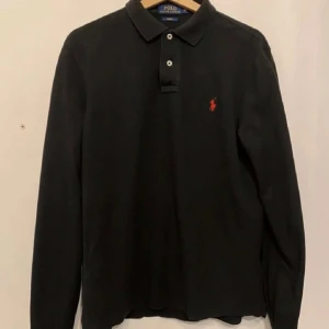 Svart pikétröja från Ralph Lauren - Säljer en stilren svart pikétröja från Ralph Lauren. Den har långa ärmar och en klassisk krage med knappar. Perfekt för både vardag och lite finare tillfällen. Den röda loggan på bröstet ger en snygg kontrast. Passar bra till jeans eller chinos!