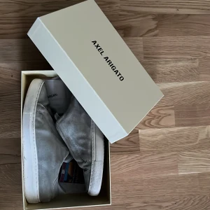 Axel Arigato Skor - Säljer ett par grå sneakers från Axel Arigato i mocka. De har en stilren design med dragkedja framtill istället för traditionell snörning. Perfekta för både vardag och festliga tillfällen. Skorna är i välanvänt skick men fortfarande snygga och bekväma.