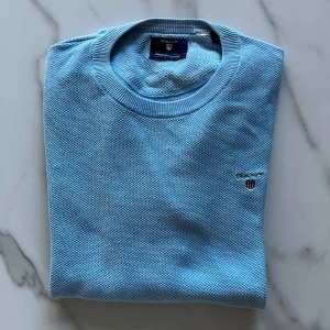 Ljusblå tröja från Gant - Säljer en stilren ljusblå tröja från Gant med rund hals och broderad logotyp på bröstet. Tröjan har en texturerad yta och långa ärmar med ribbade muddar. Perfekt för en avslappnad och snygg look.