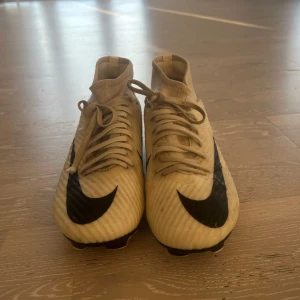 Beige Nike mercurial fotbollsskor - Snygga beige fotbollsskor från Nike med svart swoosh och snörning. Skorna har en hög krage för extra stöd och är designade för optimal prestanda på planen.