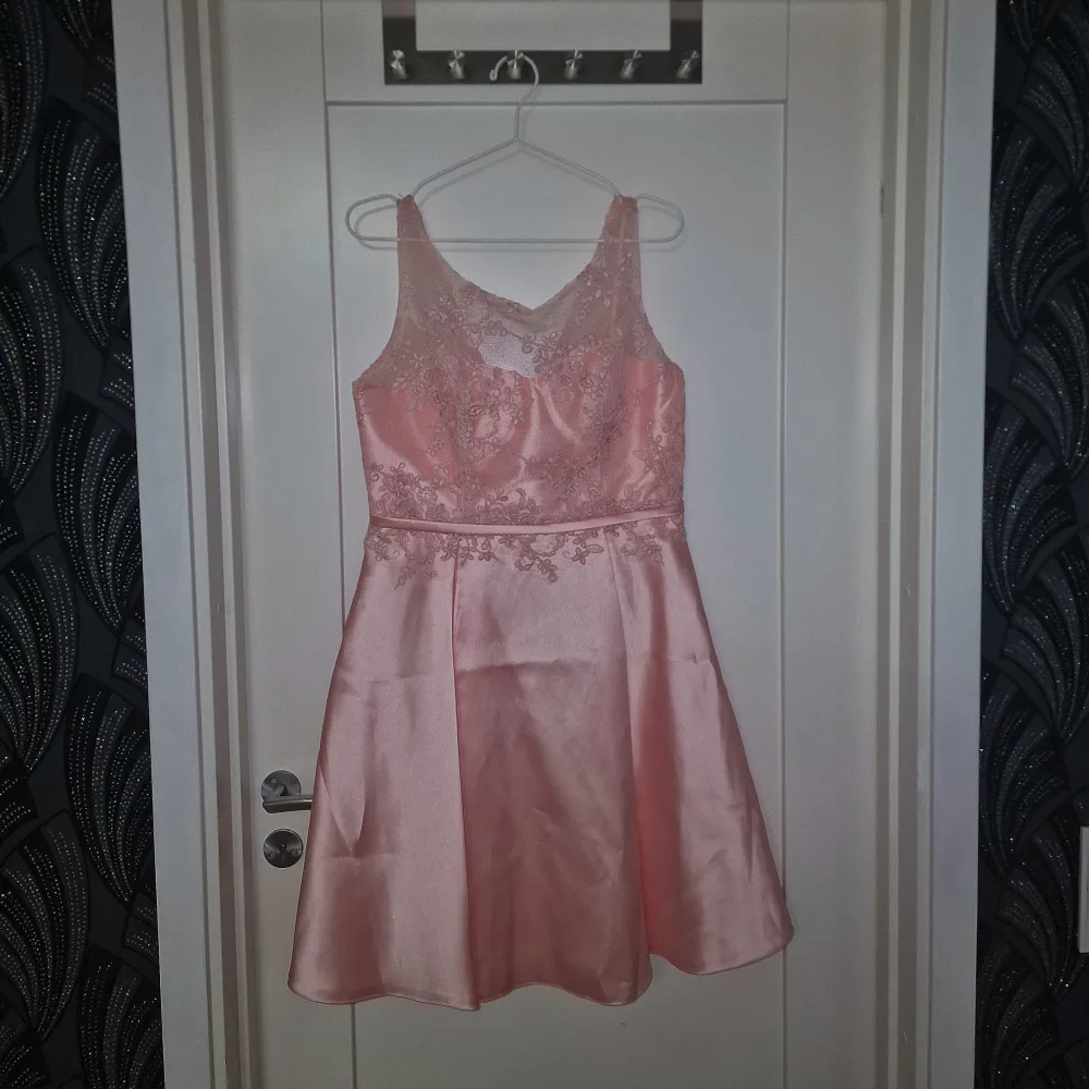 Charmig rosa klänning i satin med vackra spetsdetaljer och en elegant dragkedja i ryggen. Klänningen har en ärmlös design och en smickrande passform som passar perfekt för speciella tillfällen.. Mekot.