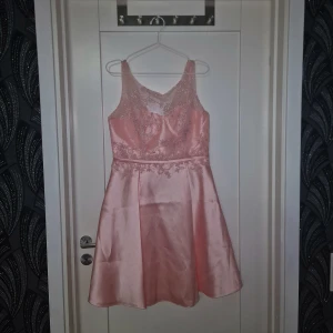 Rosa klänning med spetsdetaljer - Charmig rosa klänning i satin med vackra spetsdetaljer och en elegant dragkedja i ryggen. Klänningen har en ärmlös design och en smickrande passform som passar perfekt för speciella tillfällen.