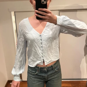 Ljusblå blus med knappar - Säljer en stilren ljusblå blus med v-ringning och knappar framtill. Blusen har långa ärmar med lätt puff och är perfekt för en elegant look.