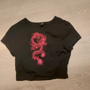Svart croppad t-shirt med draktryck - Snygg svart croppad t-shirt med ett rött draktryck på framsidan. Perfekt för en cool och avslappnad stil. Kortärmad och bekväm passform.