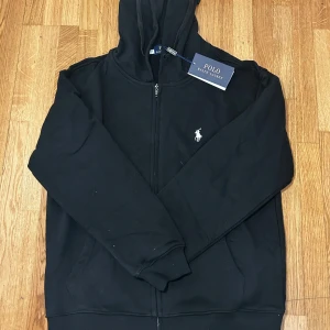 Ralph lauren zip - Säljer en stilren svart hoodie från Polo Ralph Lauren med dragkedja och det klassiska logotypbroderiet på bröstet. Perfekt för en avslappnad look. Hoodien har långa ärmar och en bekväm passform.