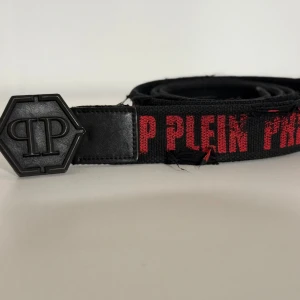 Svart bälte från Philipp Plein -  svart bälte från Philipp Plein med röd text och ett hexagonal spänne. Man må tro bältet är slitet men det är inte fallet, modellen är konstruerad på det sättet