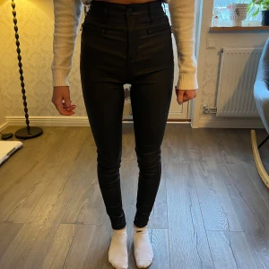 Svarta högmidjade byxor från Trend - Snygga svarta högmidjade byxor från Trend i en tight passform. Byxorna har dragkedja och knappar framtill och är perfekta för en stilren look. De är tillverkade i ett syntetmaterial som ger en glansig finish.